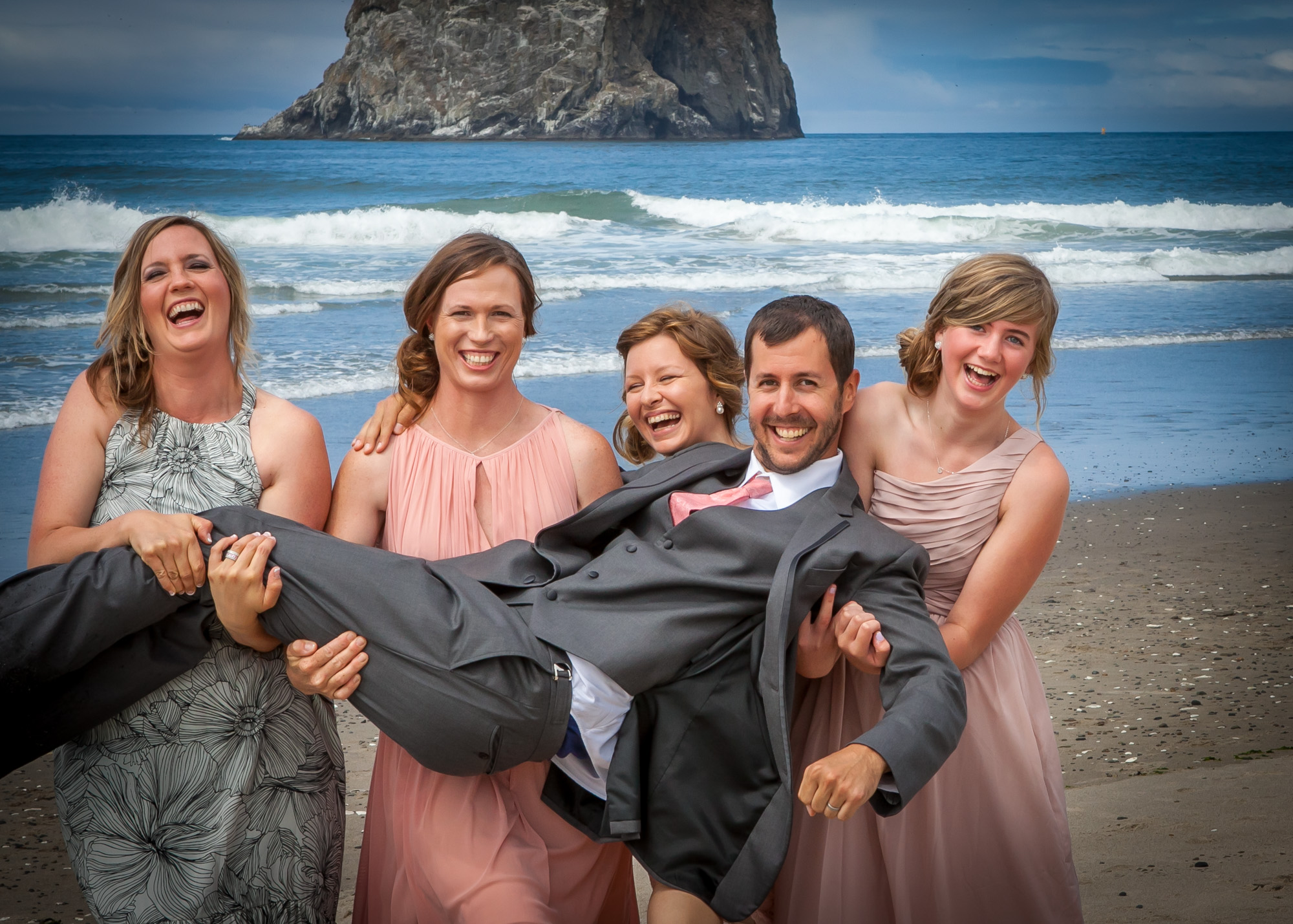 Groom Carried on the Shoulders of Laughing Bridesmaids on the Beach, With Ocean Waves and a Sea Stack in the Background.