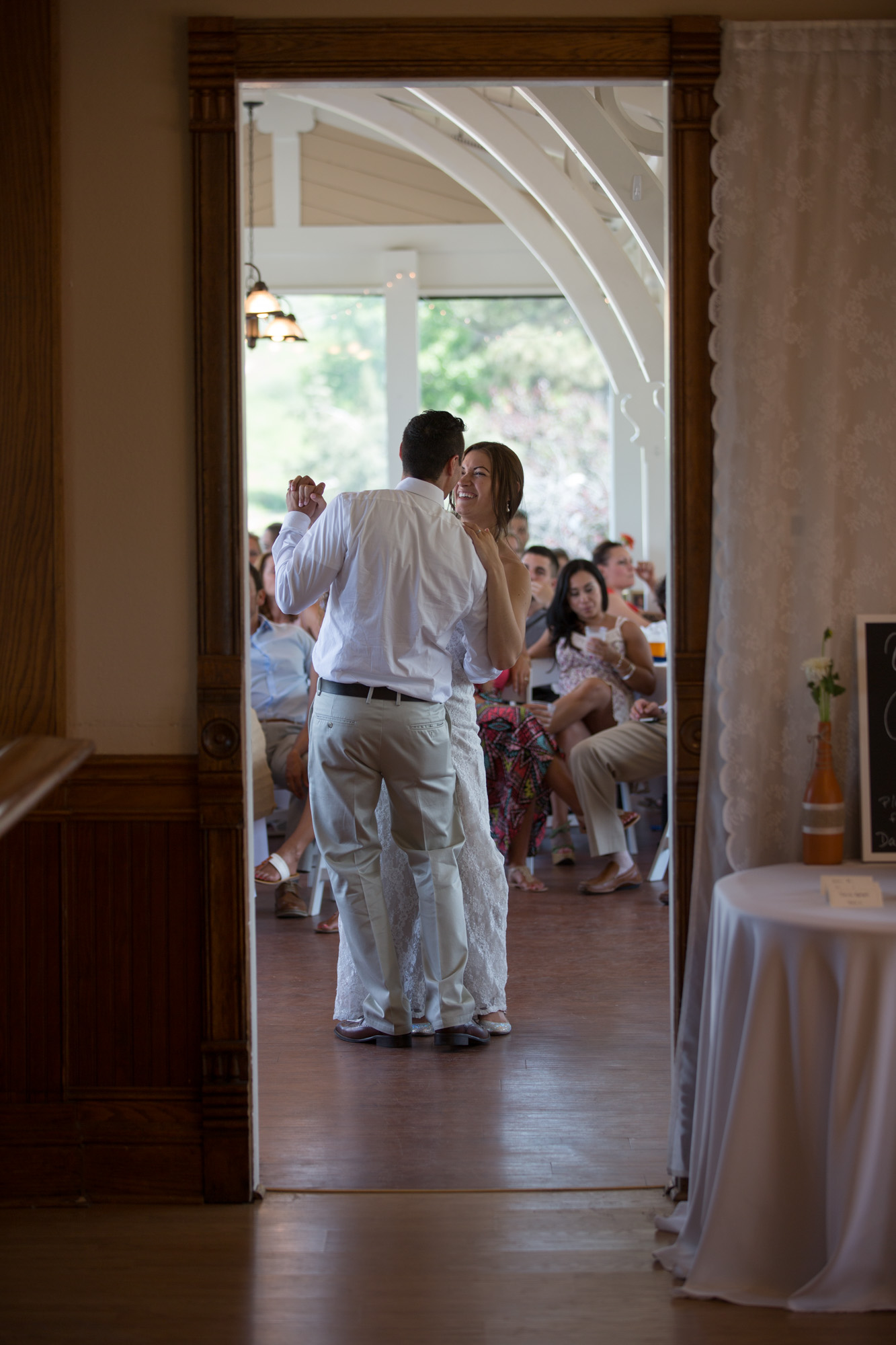 Bride and groom sharing their first dance, viewed through a doorway, with guests watching in the background.