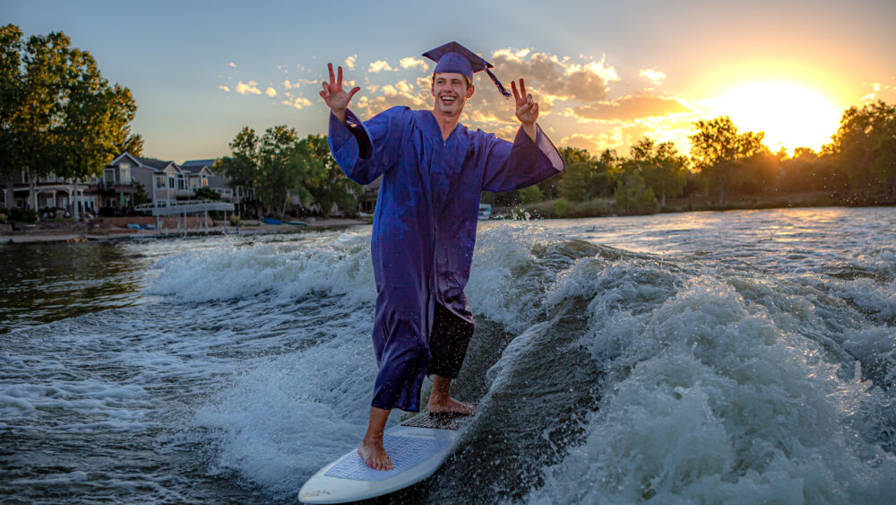 Joyful senior graduate surfing on a wave in full graduation attire during a vibrant sunset